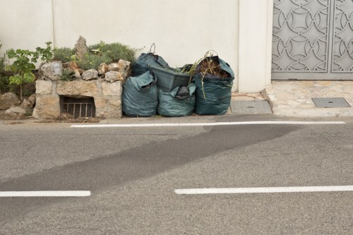 The image shows three large dark green refuse bags filled with household or garden waste placed on the pavement near a white wall with a small garden bed containing rocks and plants to the left. The bags are leaning against or resting close to the wall, with some loose plant debris visible on top of one or more bags. In the background, part of a metal gate or fence with decorative patterns is visible on the right side. The scene is set along a paved road with white painted lines marking the edge, and the overall environment suggests a typical residential or suburban area in Chalfont Saint Giles. The presence of the waste bags indicates rubbish removal activity, likely undertaken by Commercial Waste Chalfont Saint Giles, a local rubbish clearance service. The lighting is natural, with a clear view of textures including the crumpled plastic of the bags, the roughness of the gravel, and the smooth surface of the road and wall, providing an accurate visual depiction of a waste collection scene in the local area.