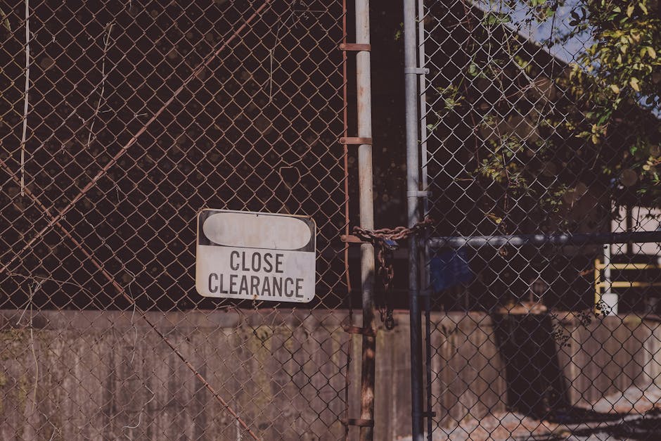 A metal chain and lock securing a double metal gate with a 'Close Clearance' sign attached, located in front of a chain-link fence that appears weathered with rust on some parts. The gate's iron bars 